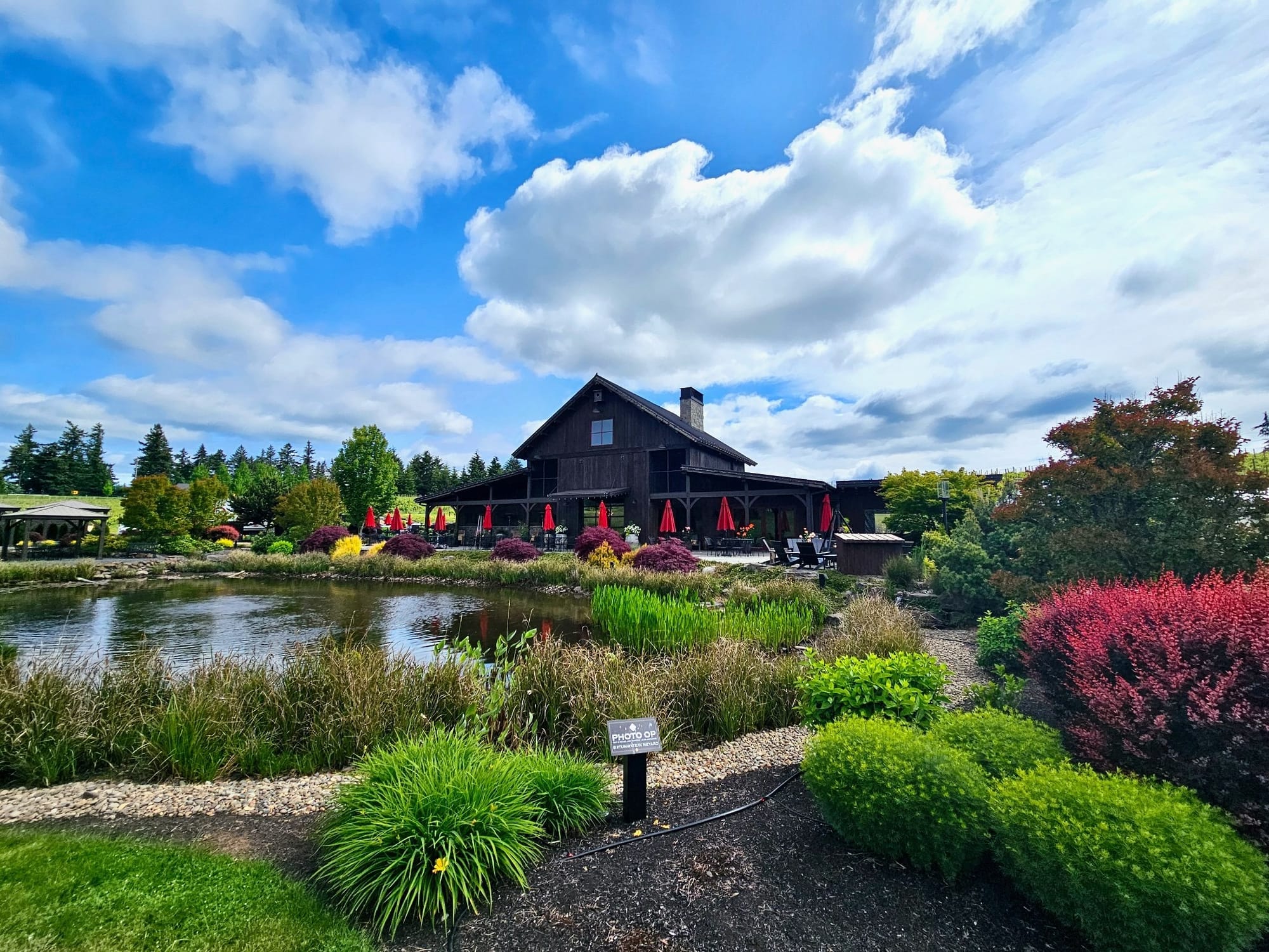 Tumwater Vineyard's barrel house tasting room in West Linn, Oregon, surrounded by a stocked trout pond, lush gardens, and outdoor seating with red umbrellas under a blue sky