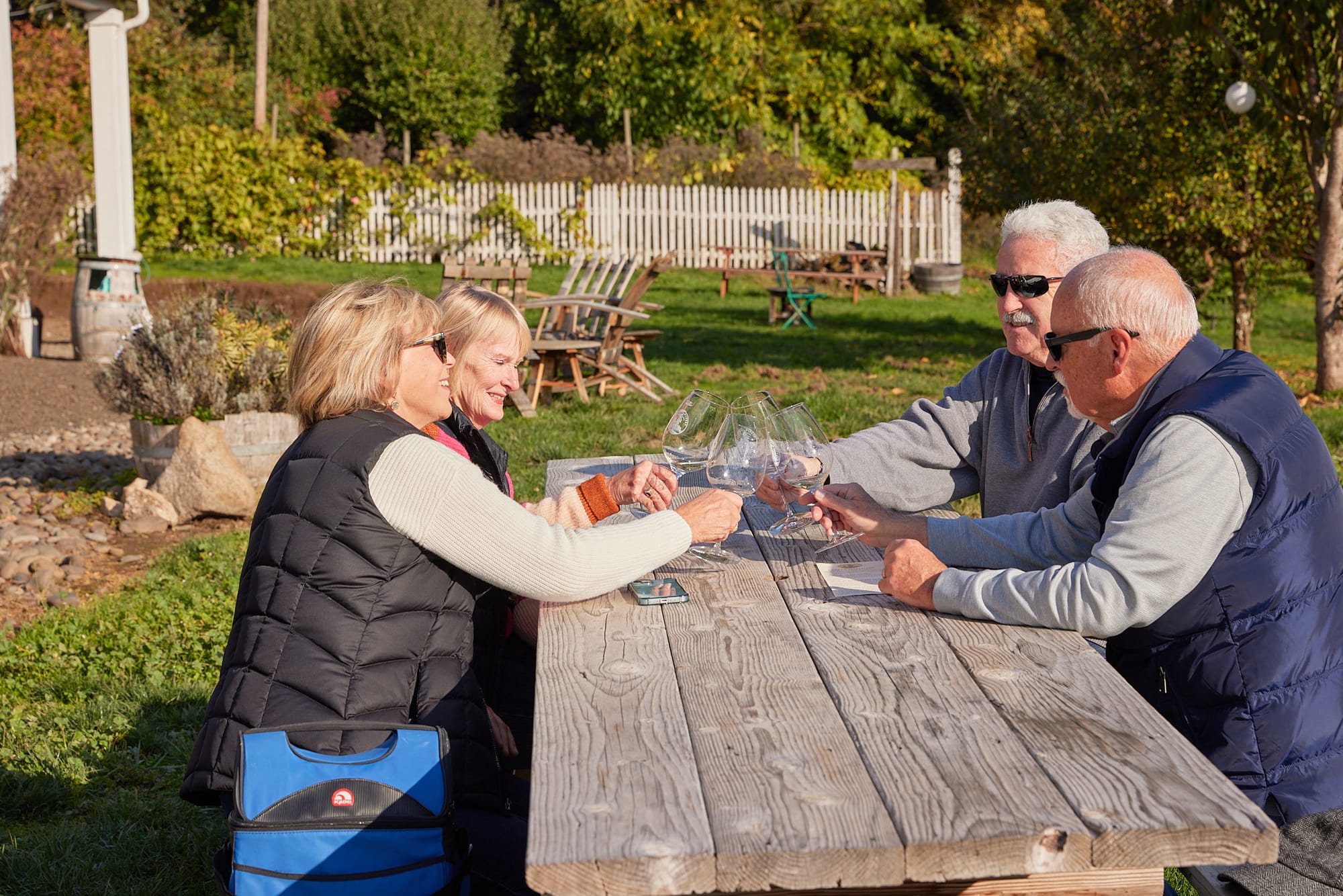 Four guests toasting with white wine glasses at an outdoor picnic table at Lady Hill Winery in St. Paul, Oregon, with vineyard rows and autumn foliage in the background on a sunny fall day