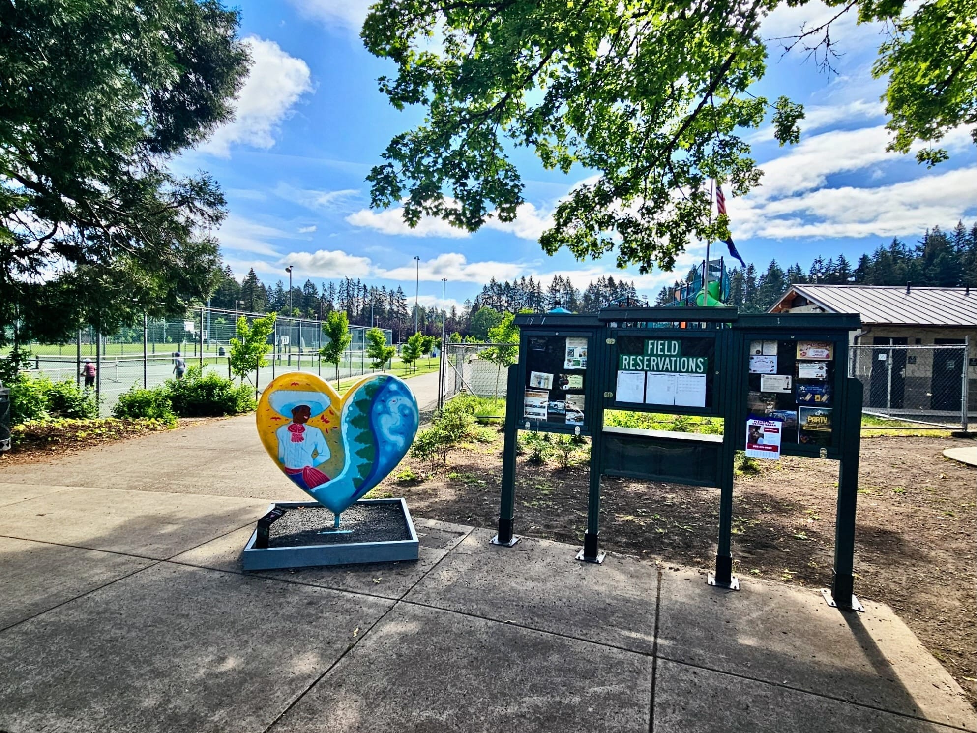 Colorful heart-shaped public art sculpture at Wilsonville Memorial Park in Oregon, with tennis courts, tall evergreen trees, and a field reservations board visible on a sunny day