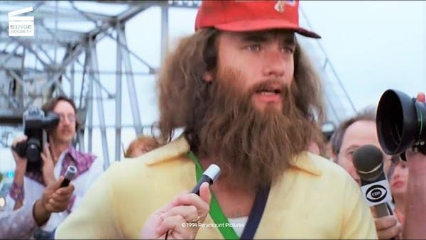 Tom Hanks as Forrest Gump. He is in a yellow polo and red hat. He has a long beard and surrounded by microphones and cameras. A bridge is behind him.
