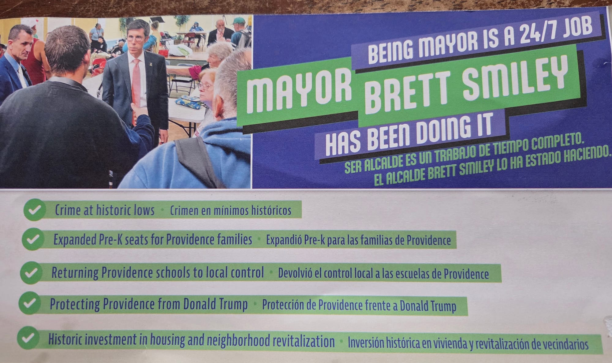 Front of mailer featuring Smiley in what appears to be a classroom. In large text, the mailer reads: “Being Mayor is a 24/7 Job. Mayor Brett Smiley Has Been Doing It,” and provides the same message in Spanish in smaller text. Below, five bullet points are given next to checkmark symbols, which read: “Crime at historic lows; Expanded Pre-K seats for Providence families; Returning Providence schools to local control; Protecting Providence from Donald Trump; Historic investment in housing and neighborhood revitalization”, next to Spanish translations. (The term “vecindario”&nbsp;is used to mean “neighborhood”, which strikes me as possibly nonstandard, but I am not fluent.) Almost all Spanish text on this and other mailpieces is smaller than the English text.