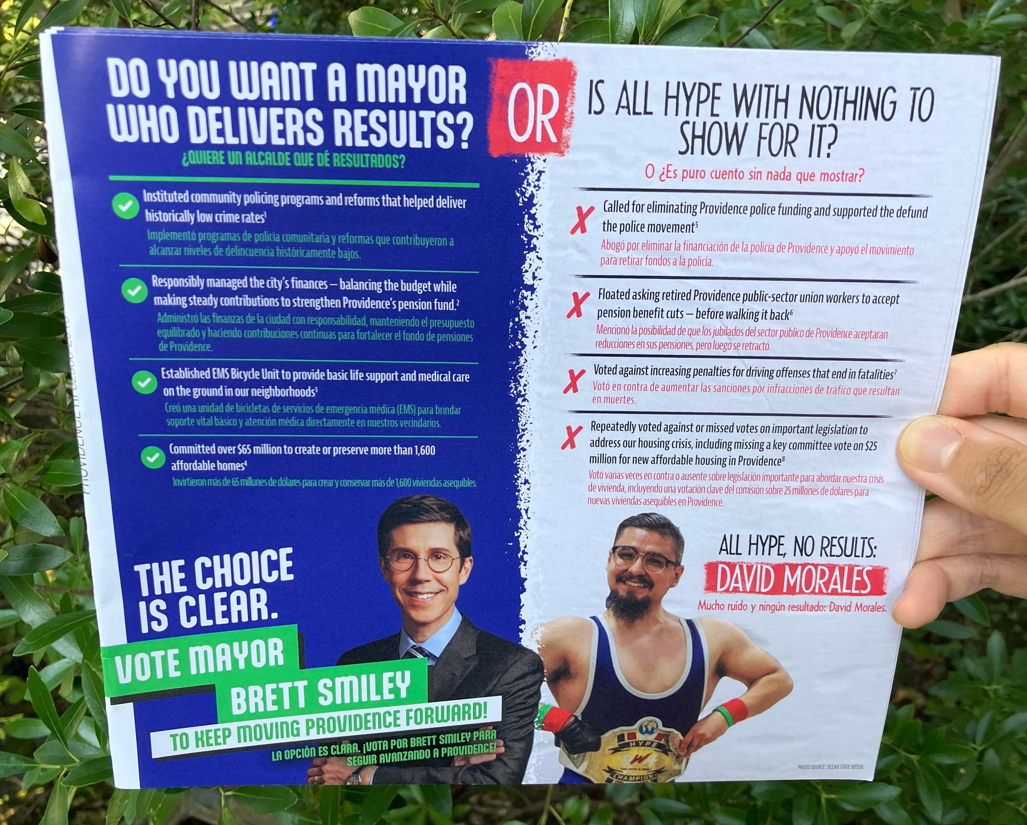 One side of a bilingual contrast mailpiece. At top left, on blue background, it says “Do you want a mayor who delivers results?” then in large letters “or” and at top right “is all hype with nothing to show for it?” On the left side are four pro-Smiley points, each next to a green check mark, asserting Smiley has “helped deliver historically low crime rates”, “responsibly managed the city's finances”, “established [an] EMS Bicycle Unit”, and “committed over $65 million to create or preserve more than 1,600 affordable homes”. At right, four anti-Morales points appear next to red X marks, asserting Morales “supported the defund the police movement”, “floated […] public-sector union […] pension benefit cuts”, “voted against increasing penalties for driving offenses that end in fatalities”, and “repeatedly voted against or missed important legislation to address our housing crisis”. At bottom left, it says “The Choice Is Clear: Vote Mayor Brett Smiley to Keep Moving Providence Forward!”&nbsp;next to a photo of Smiley in a suit. At bottom right, it says “All Hype, No Results: David Morales”, next to an image of Morales's head attached to a posing muscular figure with a WWE-style belt reading “Hype Champion”. If you'd like a full transcription of all text, contact me and I'll get it to you.