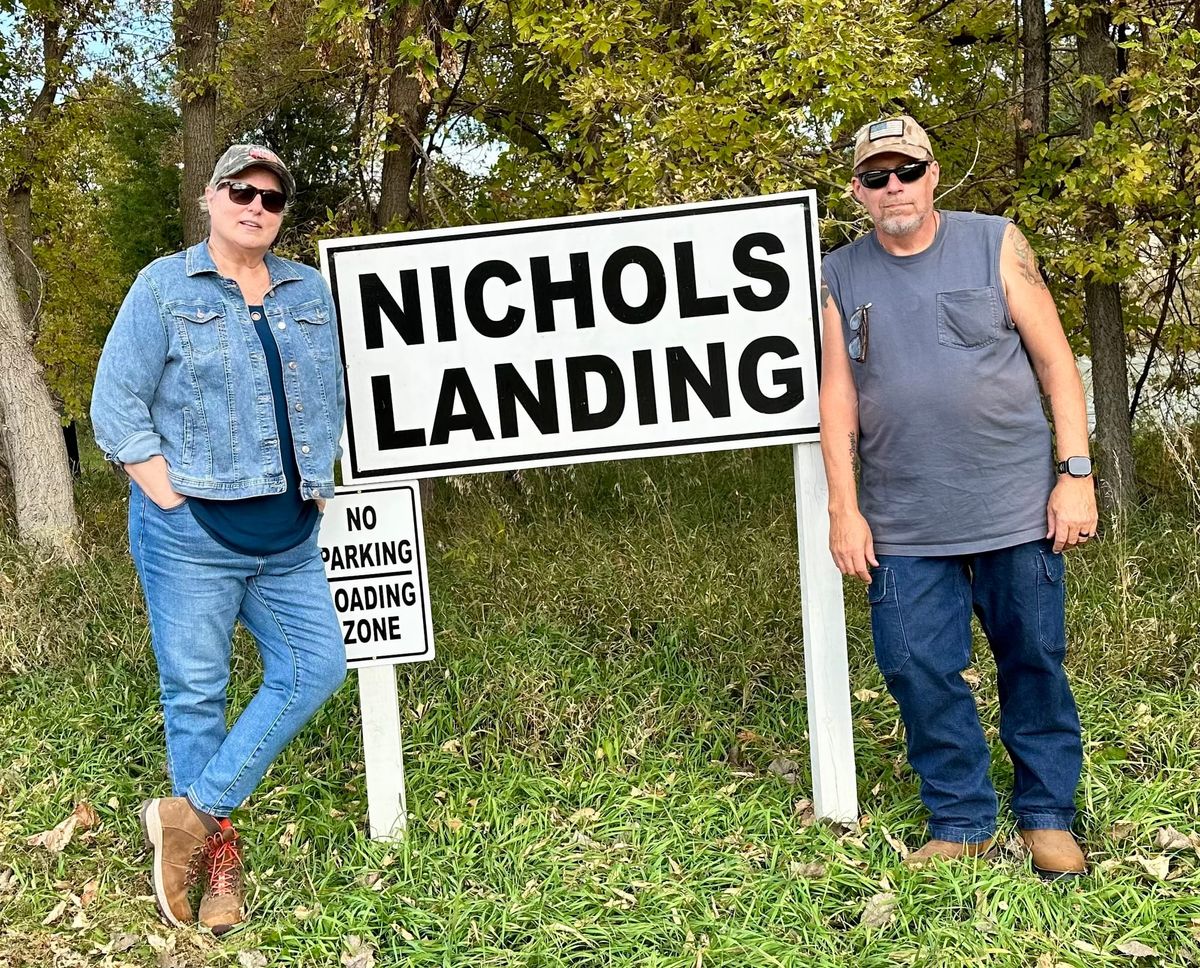 Phil and Lisa standing on either side of a white sign reading “Nichols Landing” near Smith Falls State Park in Nebraska.