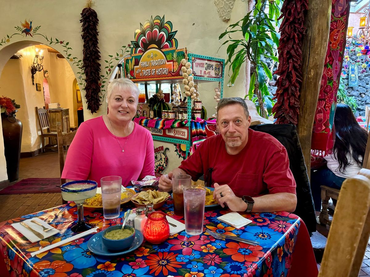 Phil and Lisa seated at a colorful table inside La Posta de Mesilla, surrounded by adobe walls, hanging chile ristras, and Southwestern décor.