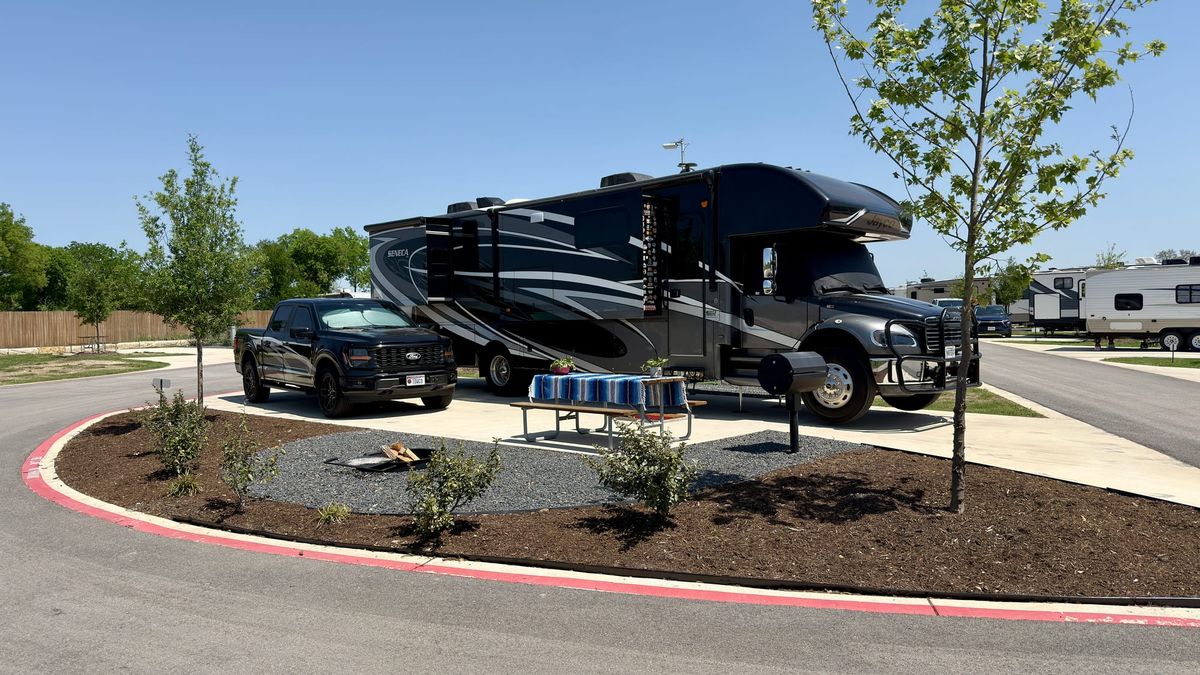 Harriet, a large Super C motorhome, parked beside Vern on a wide concrete RV site at Gather Campground in Belton, Texas, with a picnic table, fire ring, grass and young trees.