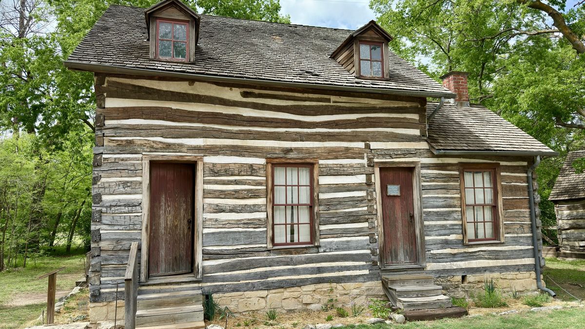 Historic two-story log house with white chinking, wooden doors and dormer windows surrounded by trees at Old Cowtown Museum in Wichita, Kansas.