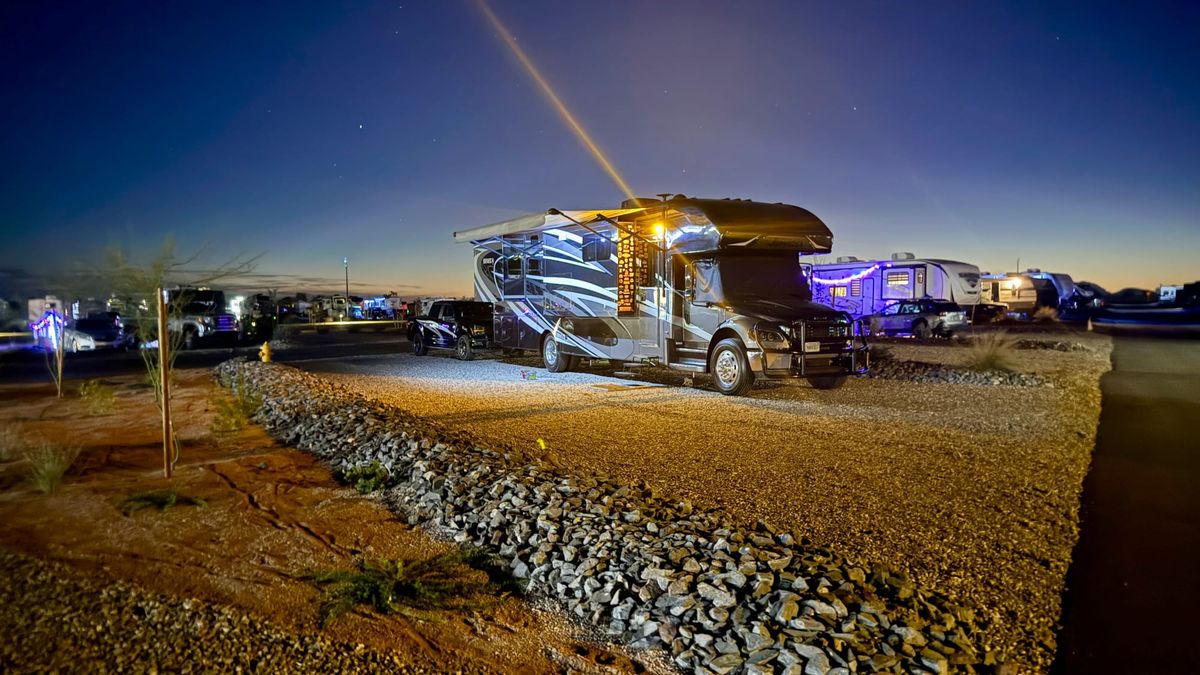 Motorhome with its awning extended and exterior lights on at a gravel campsite at dusk.