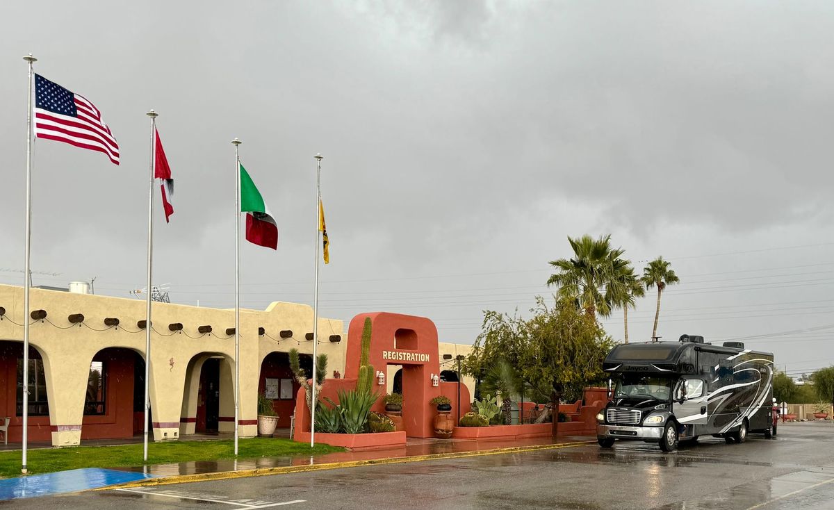 Harriet, a large Super C motorhome, parked near the registration building at Tucson KOA Resort on a rainy day, with several flags flying overhead.