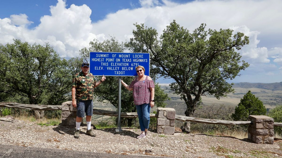 Two visitors beside the Mount Locke summit sign, with the Davis Mountains stretching behind them.