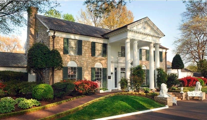 Front exterior of Graceland Mansion with its white-columned entrance, stone walls, landscaped lawn, and surrounding trees.