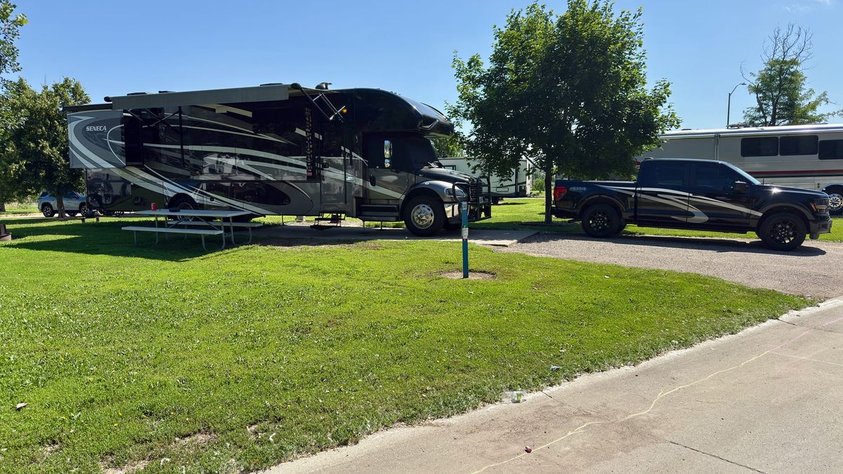 Motorhome with its awning extended beside a pickup at a grassy Scenic Park campsite, with neighboring RVs and shade trees.