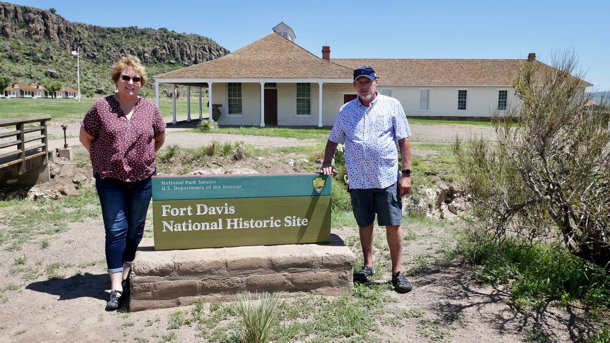 Two visitors standing beside the Fort Davis National Historic Site sign, with a historic white building and rocky hillside in the background.