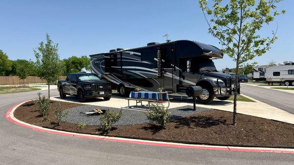 Harriet, a large Super C motorhome, parked beside Vern on a wide concrete RV site at Gather Campground in Belton, Texas, with a picnic table, fire ring, grass and young trees.