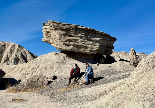 Two visitors sitting beneath a broad, overhanging rock formation at Toadstool Geologic Park in Nebraska.