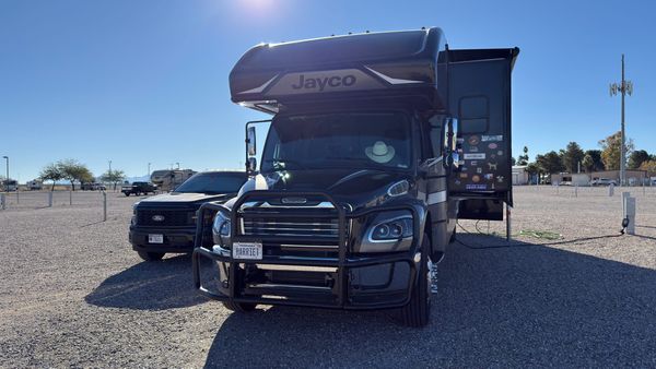 Harriet, a large Jayco Super C motorhome, parked with Vern beside it on a spacious gravel RV site at the Pima County Fairgrounds in Tucson, Arizona.