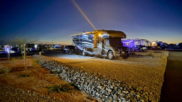 Motorhome with its awning extended and exterior lights on at a gravel campsite at dusk.