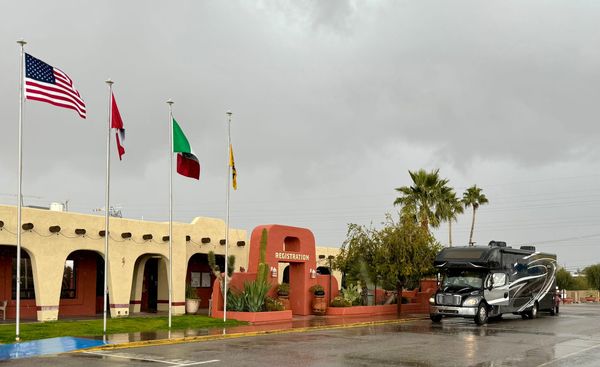 Harriet, a large Super C motorhome, parked near the registration building at Tucson KOA Resort on a rainy day, with several flags flying overhead.