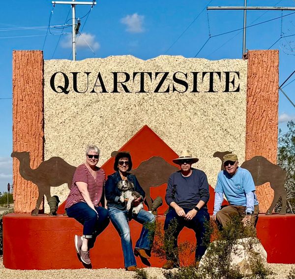 Four adults sitting in front of a large colorful Quartzsite sign decorated with camels in Quartzsite, Arizona.