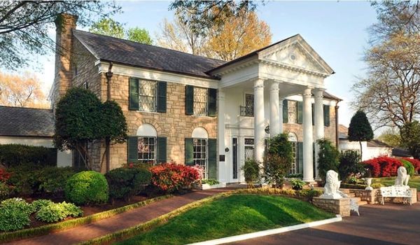 Front exterior of Graceland Mansion with its white-columned entrance, stone walls, landscaped lawn, and surrounding trees.