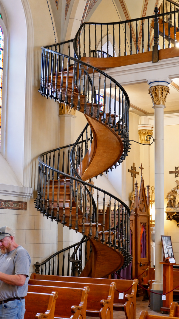 Wooden spiral staircase with two complete turns and ornate dark railings inside Loretto Chapel in Santa Fe, New Mexico. A visitor stands near the pews in the foreground.