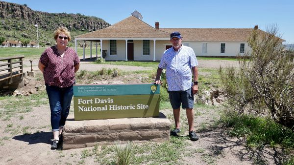 Two visitors standing beside the Fort Davis National Historic Site sign, with a historic white building and rocky hillside in the background.