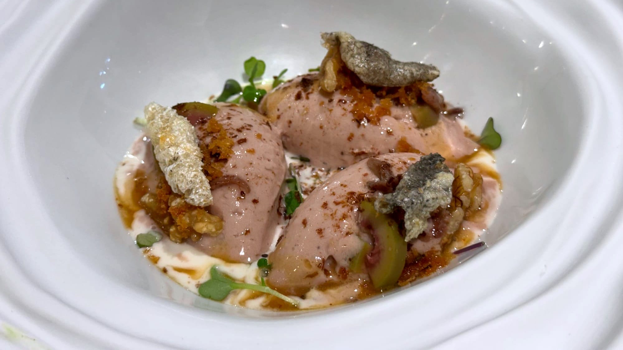 Three pink quenelles of cod liver ice cream garnished with crumbs, herbs, and crisp bits in a shallow creamy sauce.