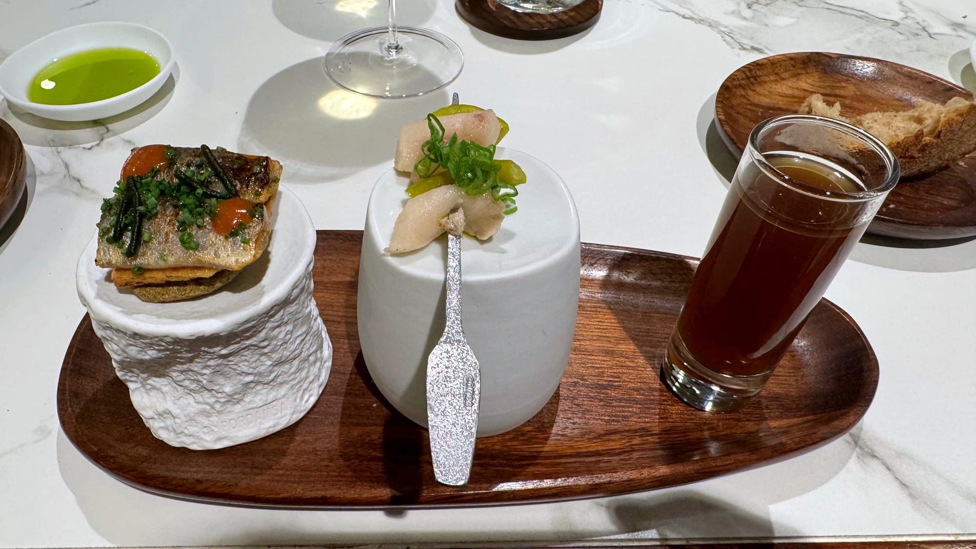 Three-part amuse on a wooden tray—sardine-topped cracker, skewer with swordfish Gilda, and a small glass of dark mushroom broth.