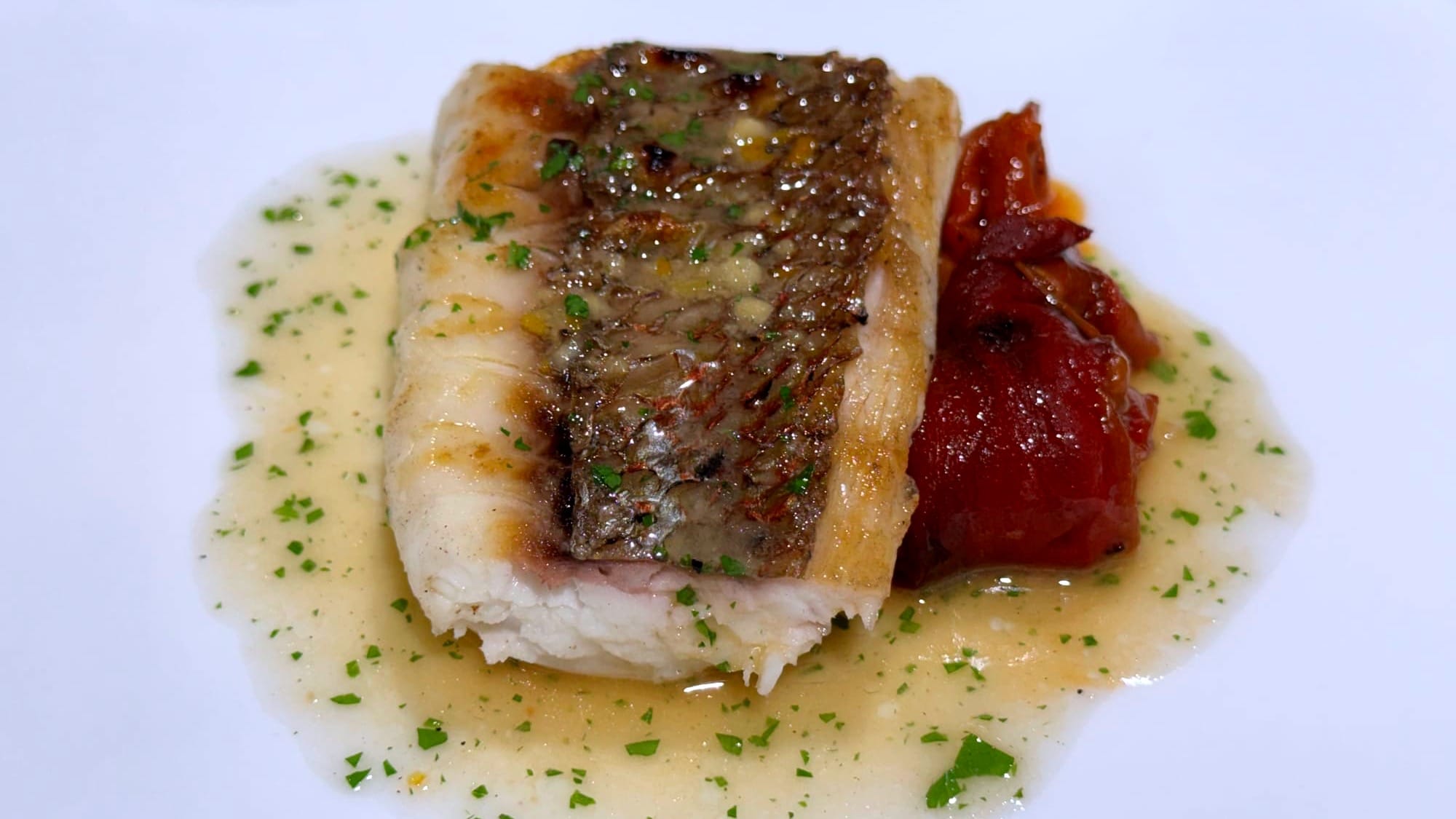 Rectangular fillet of grilled sea bream with crisp skin, parsley-speckled sauce, and roasted red peppers on a white plate.
