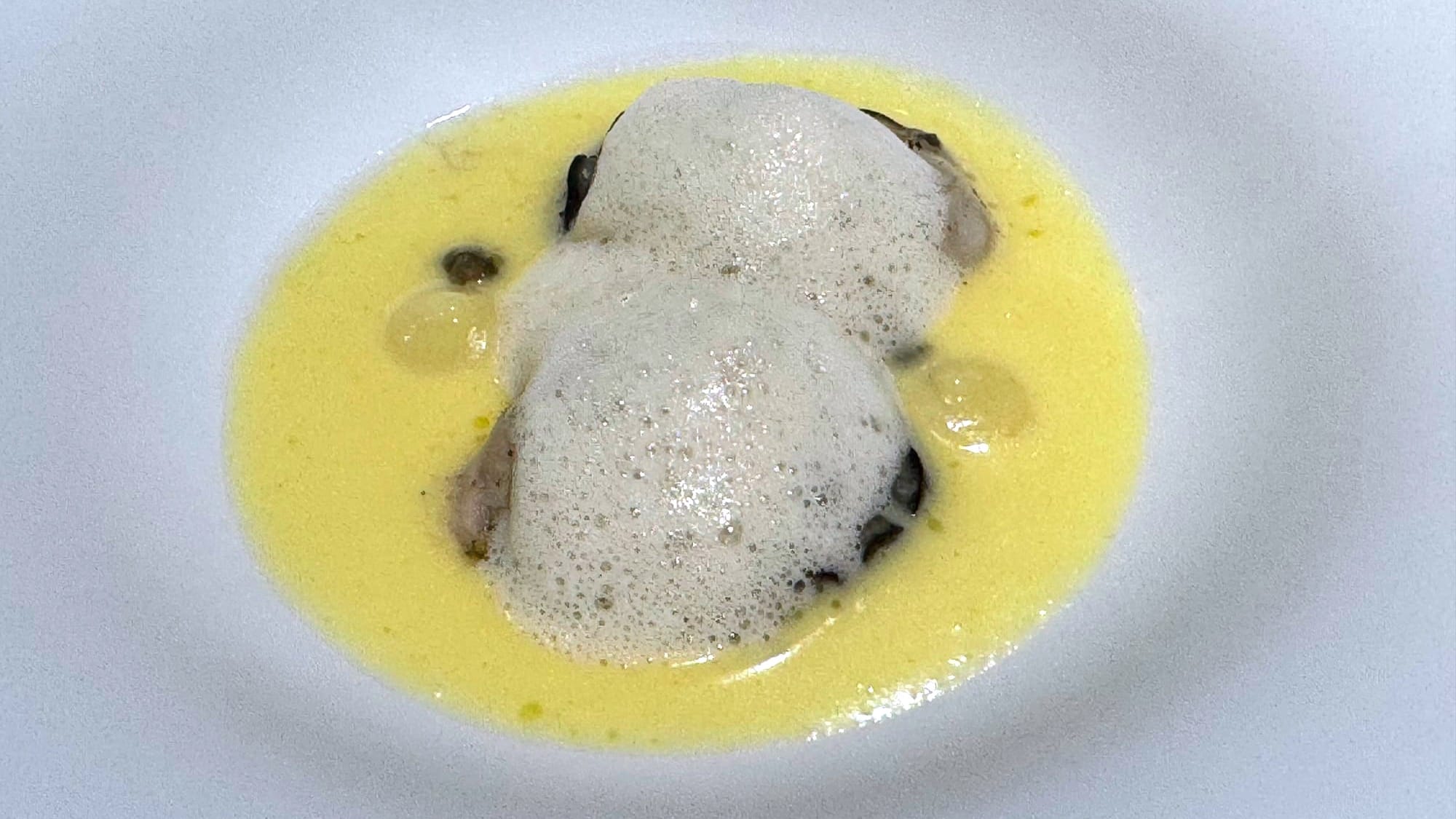 Two poached oysters under a light beurre meunière foam in a shallow yellow sauce.