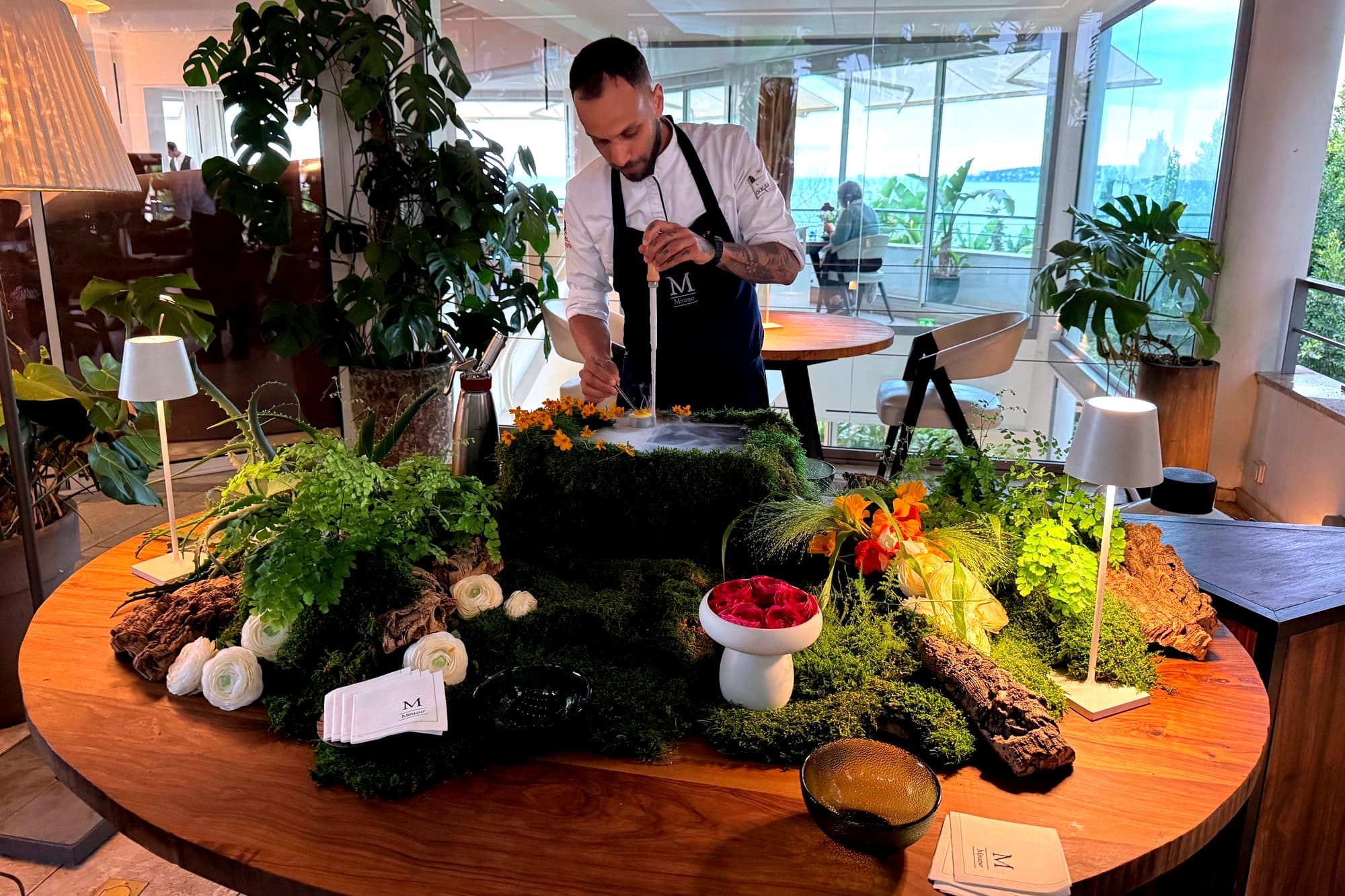 A chef preparing a solid génépi cocktail at a table decorated with moss, herbs, and flowers, arranged like a small garden, in a bright dining room with a sea view.