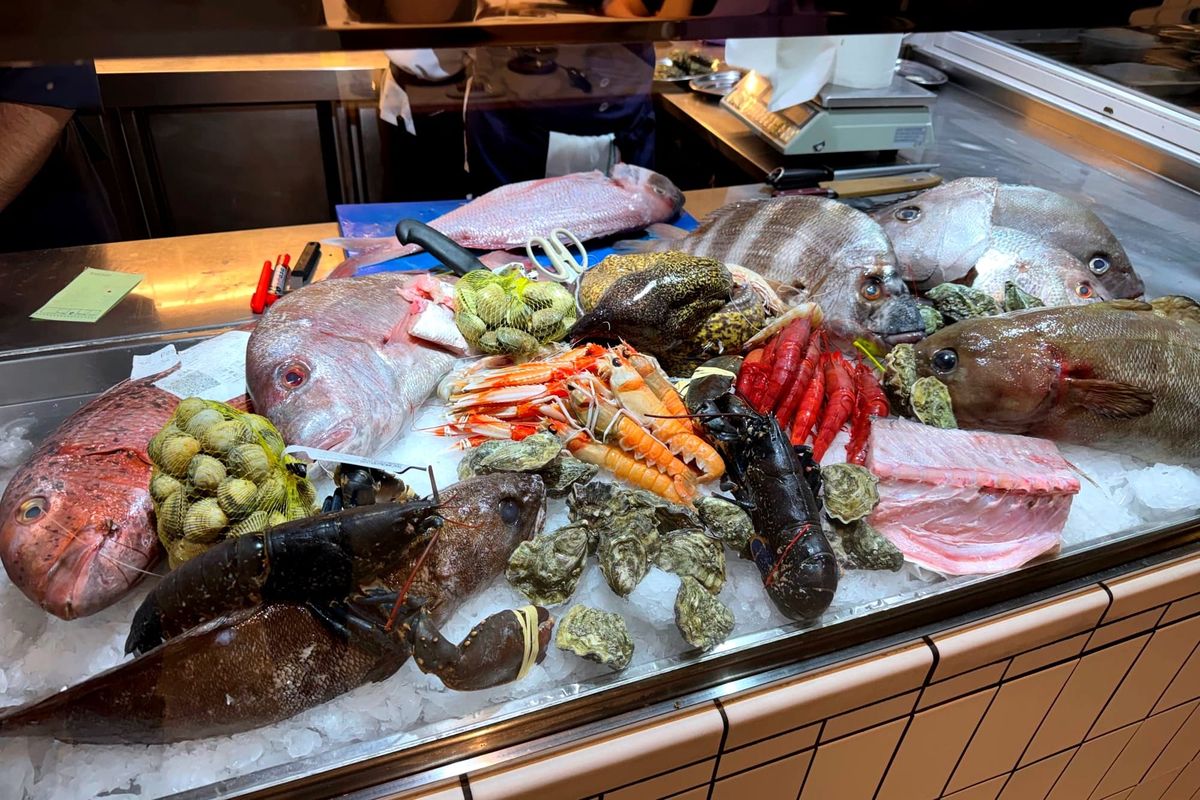 Display of whole fish, lobsters, prawns, clams, and oysters on crushed ice behind glass at the restaurant entrance.
