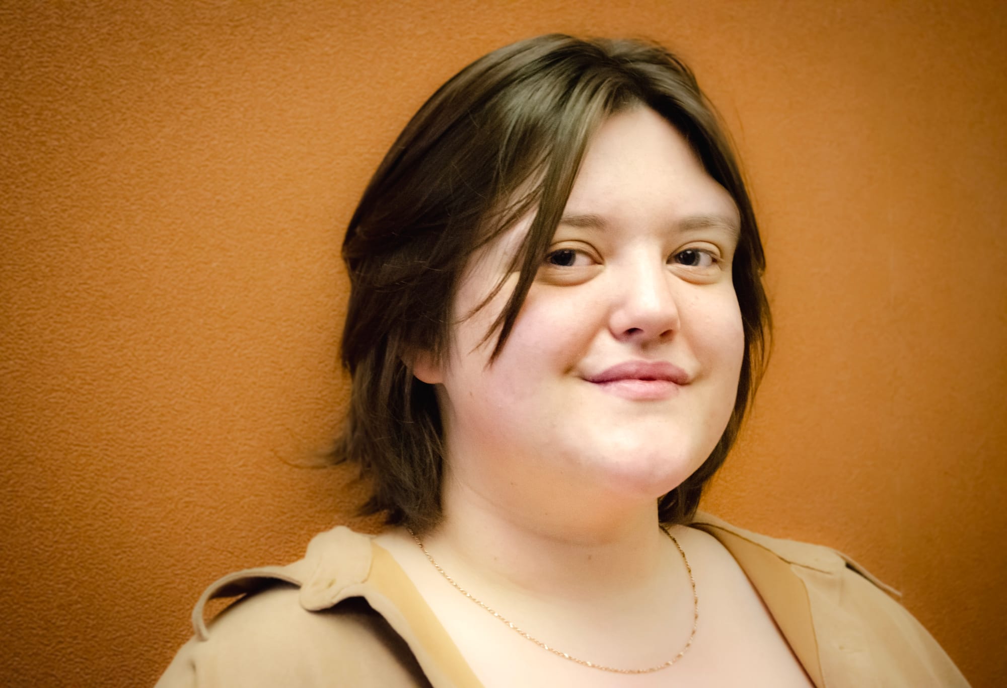 A headshot of a person with shoulder-length brown hair wearing a gold chain and a brown button up shirt against an orange background.