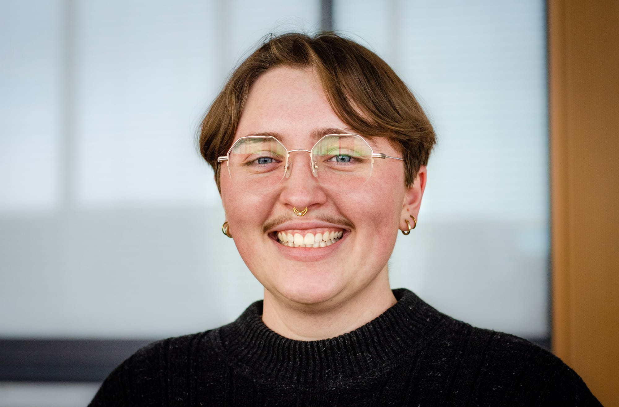 A headshot of a person with short brown hair, frameless glasses, a septum ring and wearing a black sweater.