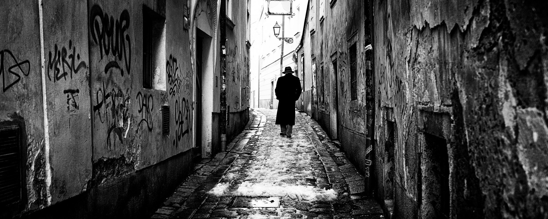 A lone figure walks down a narrow, graffiti-covered alley in a black-and-white photograph.