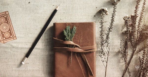Leather journal with pencil, dried herbs, and a tarot card edge, representing a 14-day tarot self-awareness practice.