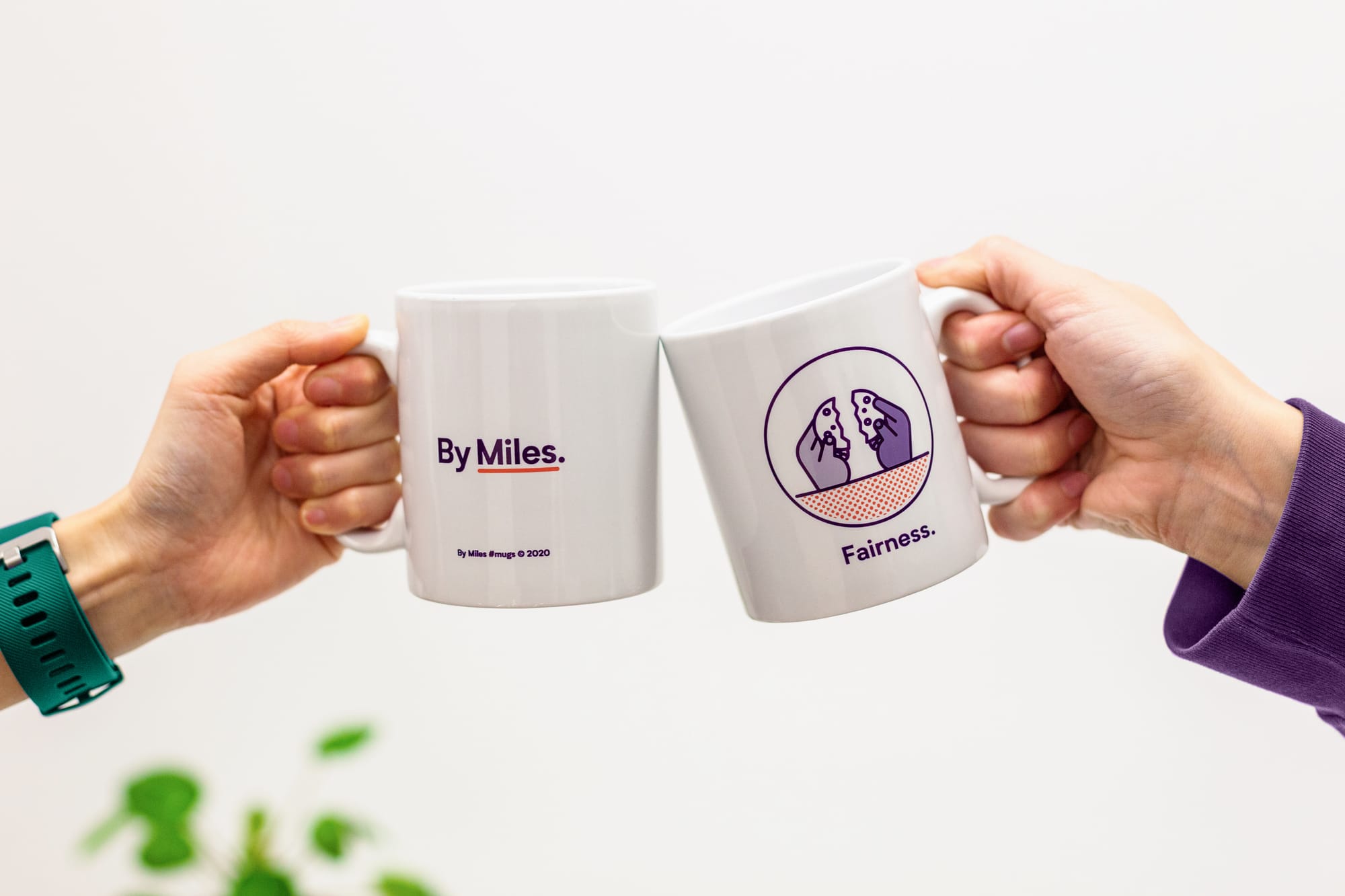 Two By Miles branded mugs clinking together, one displaying the By Miles logo, the other printed with the word Fairness, a core company value.