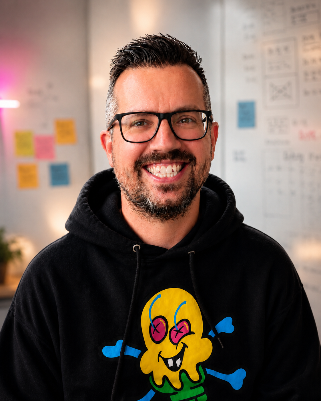 Joe Pendlebury smiling at the camera, wearing black-framed glasses and a black hoodie with a colourful skull graphic. Background shows a whiteboard covered in sticky notes and UX diagrams.