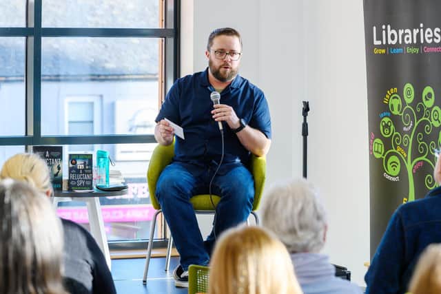 Me in a blue shirt and jeans, sitting on a low stage with a microphone, in front of an audience of library patrons