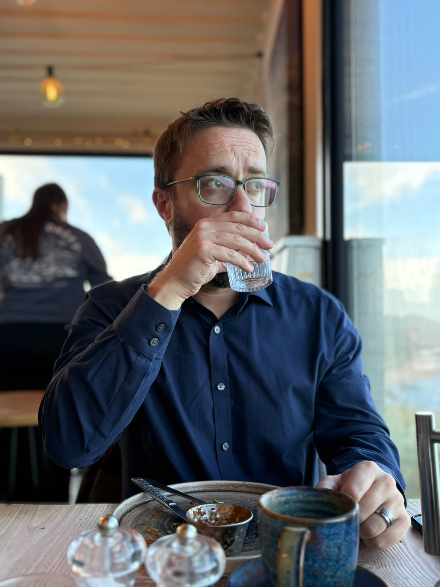 Me, in a blue shirt, drinking from a glass of water, looking out of a floor-to-ceiling window