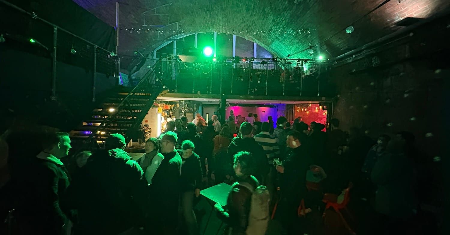 A crowded bar inside an old railway arch, lit with green light