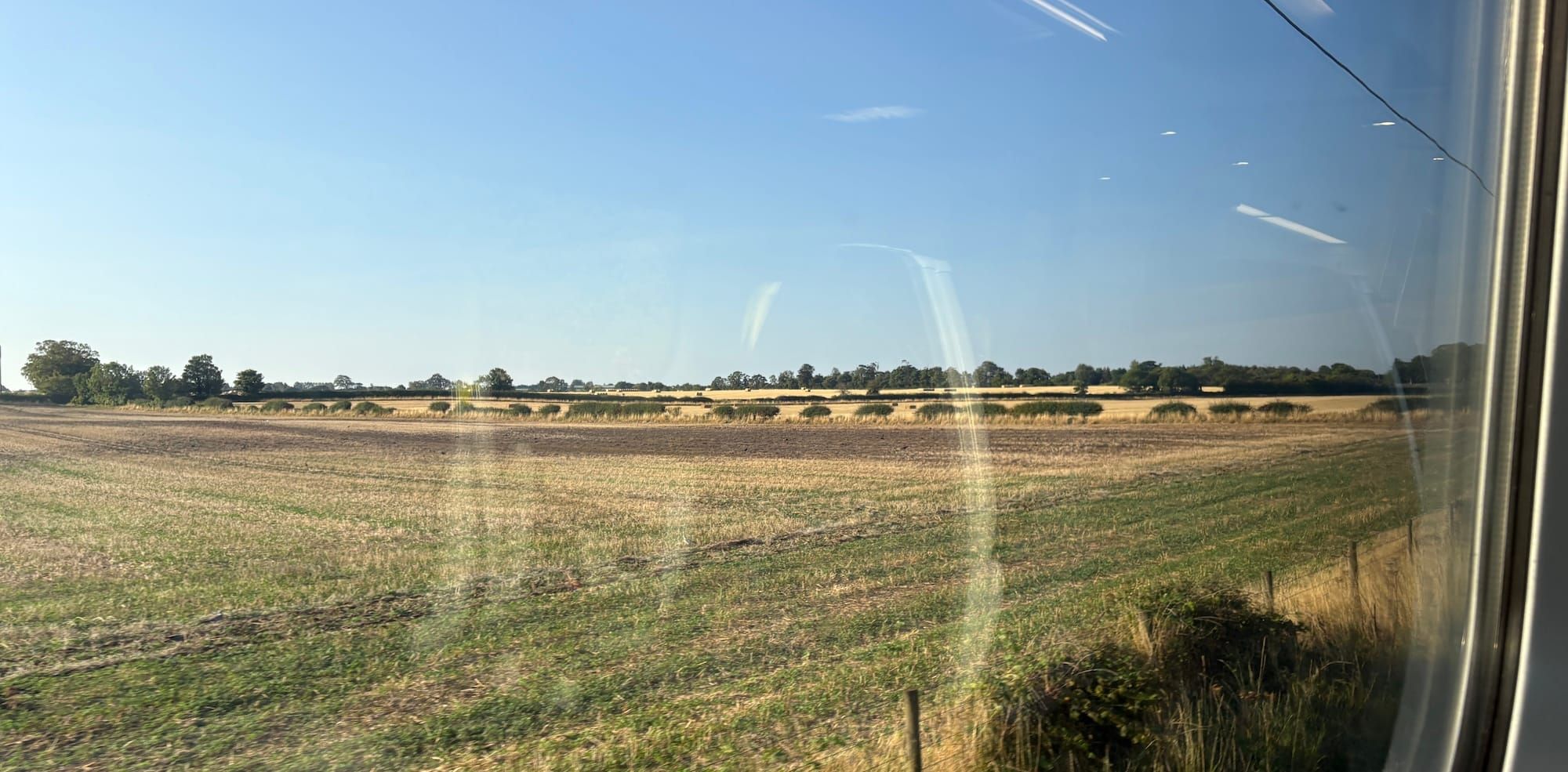 A view of fields from a train window, with trees in the distance and blue sky
