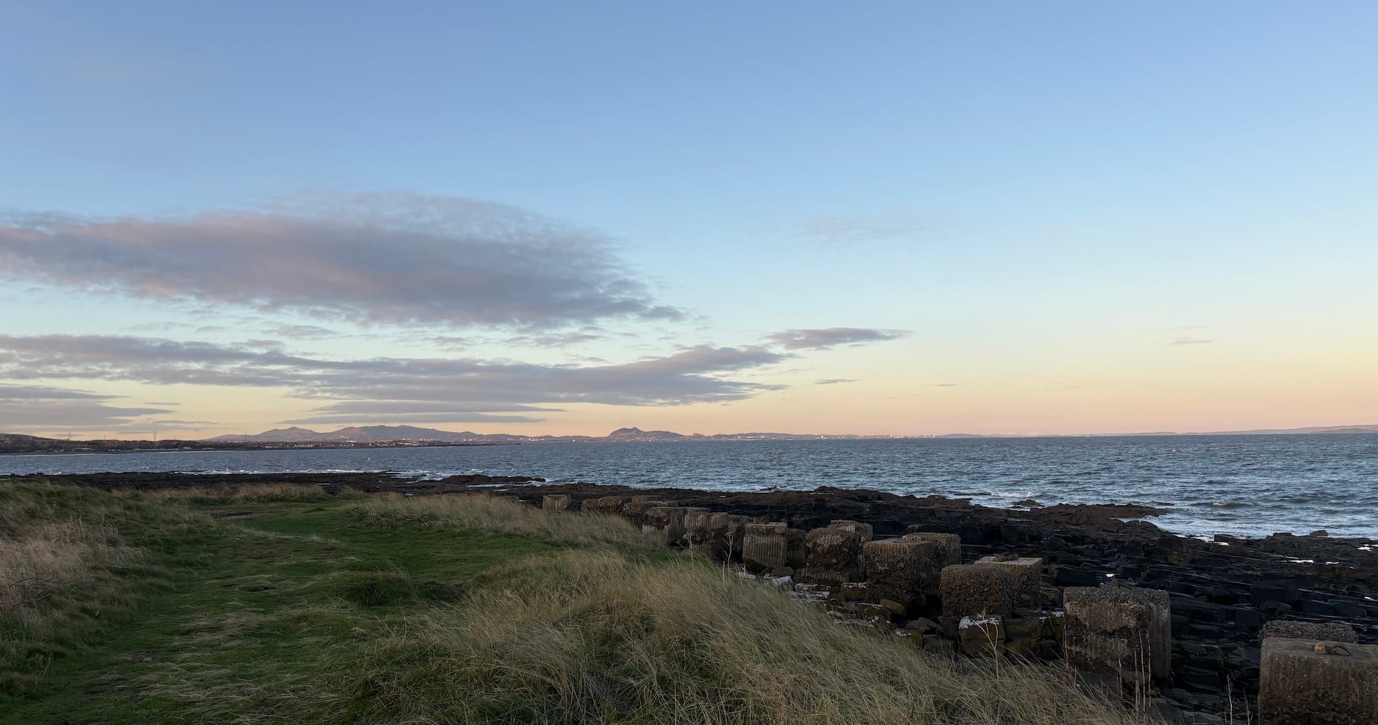 A view over the Firth of Forth to Edinburgh, with rough grass dunes, rocks and blocky anti-tank defences in the foreground