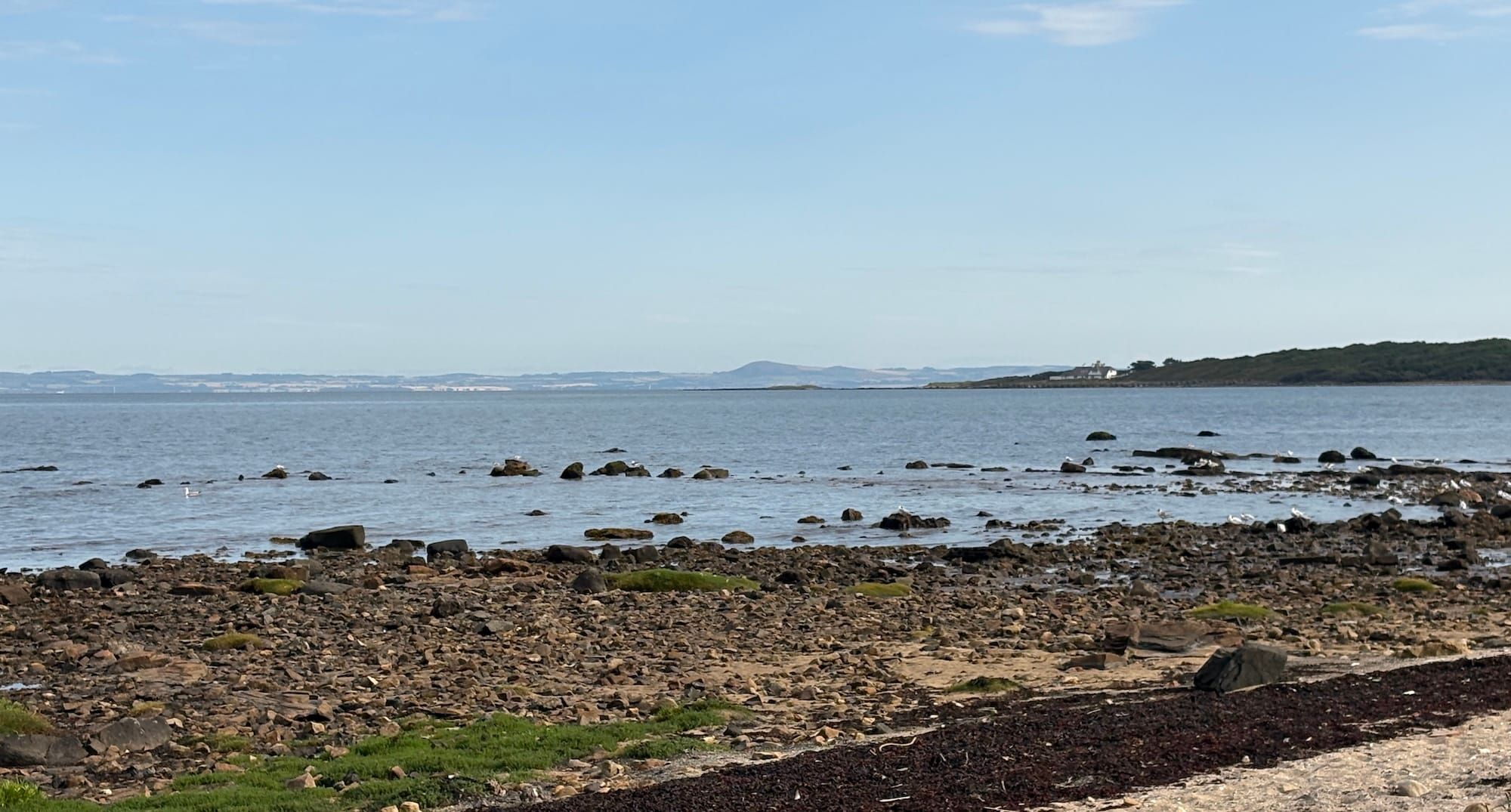Blue sky, a white house on a green, wooded point, water, rocks and sand