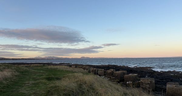 A view over the Firth of Forth to Edinburgh, with rough grass dunes, rocks and blocky anti-tank defences in the foreground