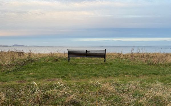A black metal bench on sand dunes, overlooking the sea, with Arthur's Seat and the Fife coast in the distance