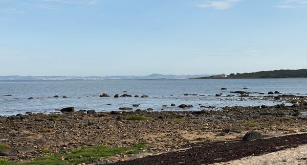Blue sky, a white house on a green, wooded point, water, rocks and sand