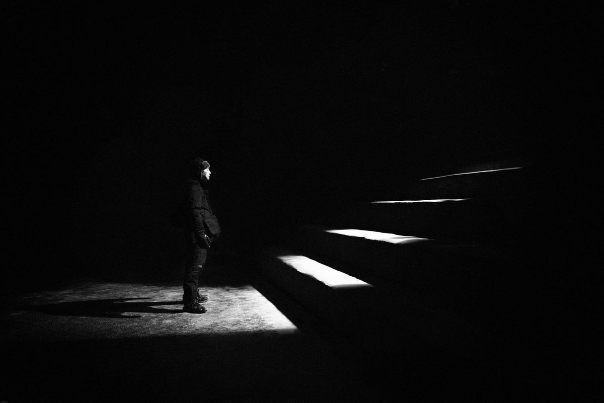 A solitary figure in a dark scene gazes at illuminated steps leading upwards, symbolizing hope or journey. Strong contrast creates a mysterious, introspective mood.