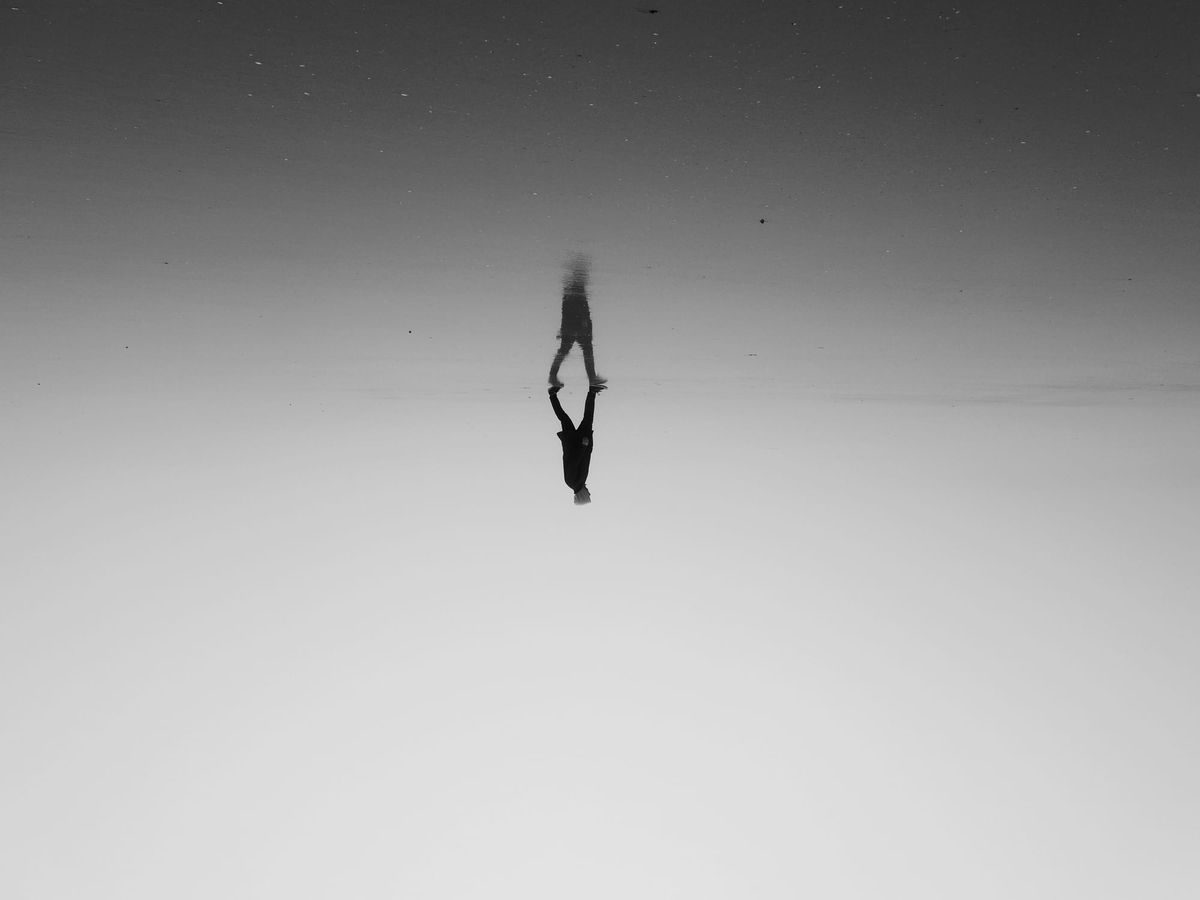 A person walks on the horizon in grayscale, reflecting upside-down in water. The scene conveys calm and surrealism with an abstract feel.