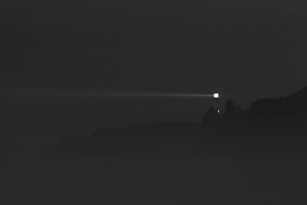 A lone lighthouse emits a bright beam across a dark, foggy landscape. The light cuts through the night, creating a sense of solitude and guidance.
