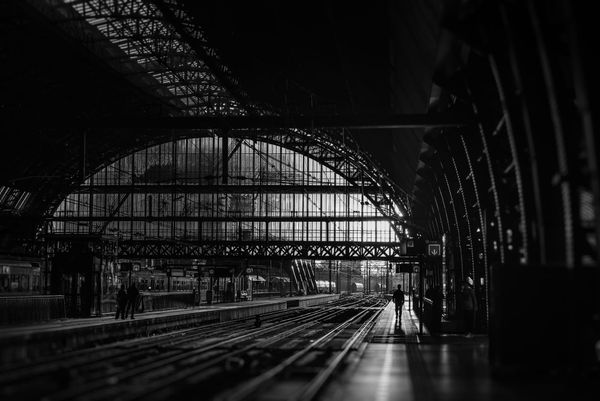 A dimly lit train station with arched glass ceilings and steel beams. Silhouetted figures walk along the platform, creating a moody, quiet atmosphere.
